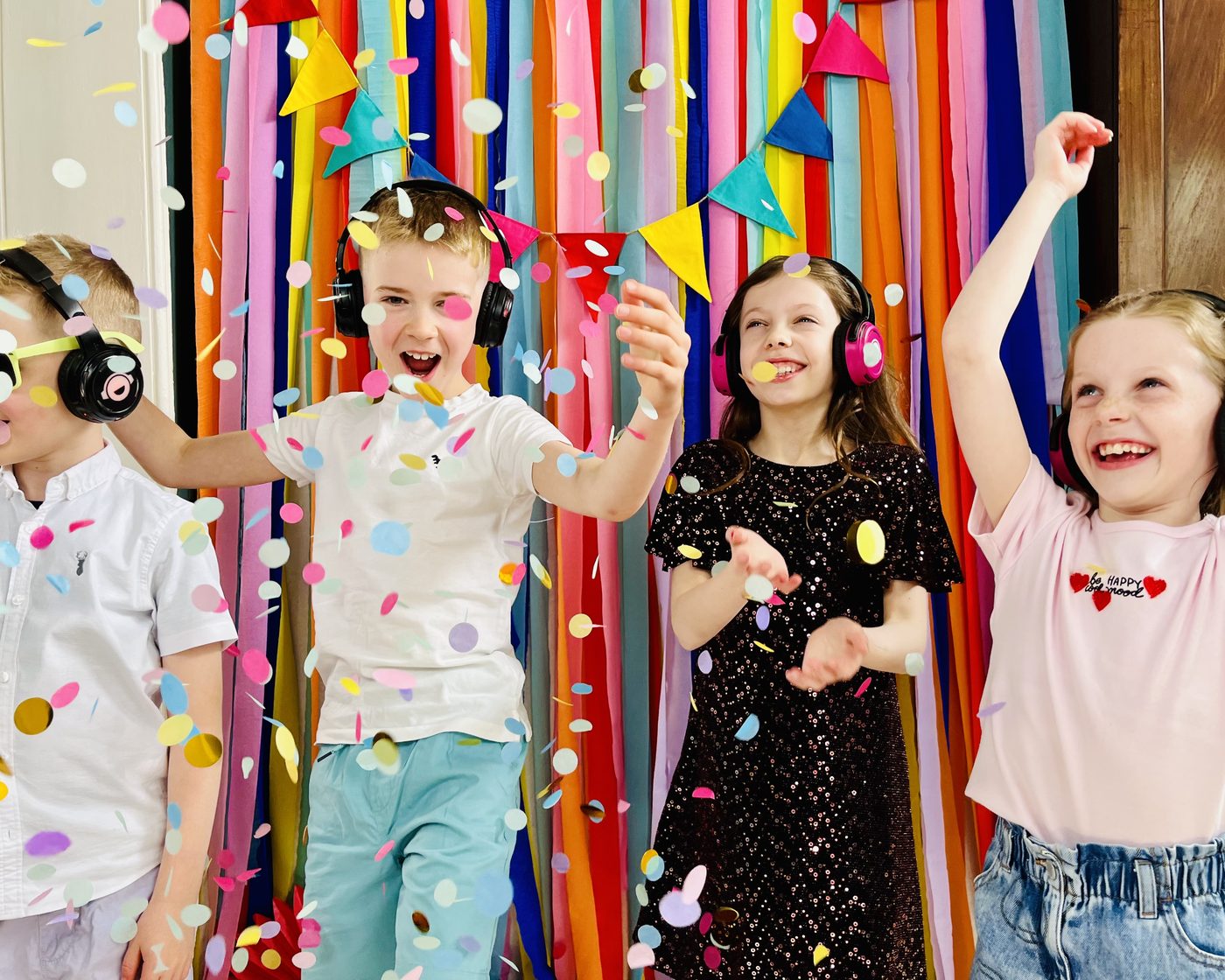 Four children laughing and dancing in colourful wireless silent disco headphones as confetti falls around them at a birthday party